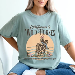 K&ouml;nnte beinhalten: Ein hellblaues T-Shirt mit einem Vintage-Grafikdesign, das ein M&auml;dchen zeigt, das ein Pferd reitet, mit dem Text "Wildflowers & Wild Horses" und "Four Fifths of Reckless and One Fifth of Jack".