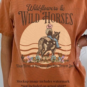 K&ouml;nnte beinhalten: Ein rostfarbenes T-Shirt mit einem Grafikdesign einer Frau, die auf einem Pferd vor einem Sonnenuntergang reitet. Der Text "Wildflowers & WILD HORSES" befindet sich oben im Design. Der Text "FOUR FIFTHS OF RECKLESS AND ONE FIFTH OF JACK" befindet sich unten im Design.