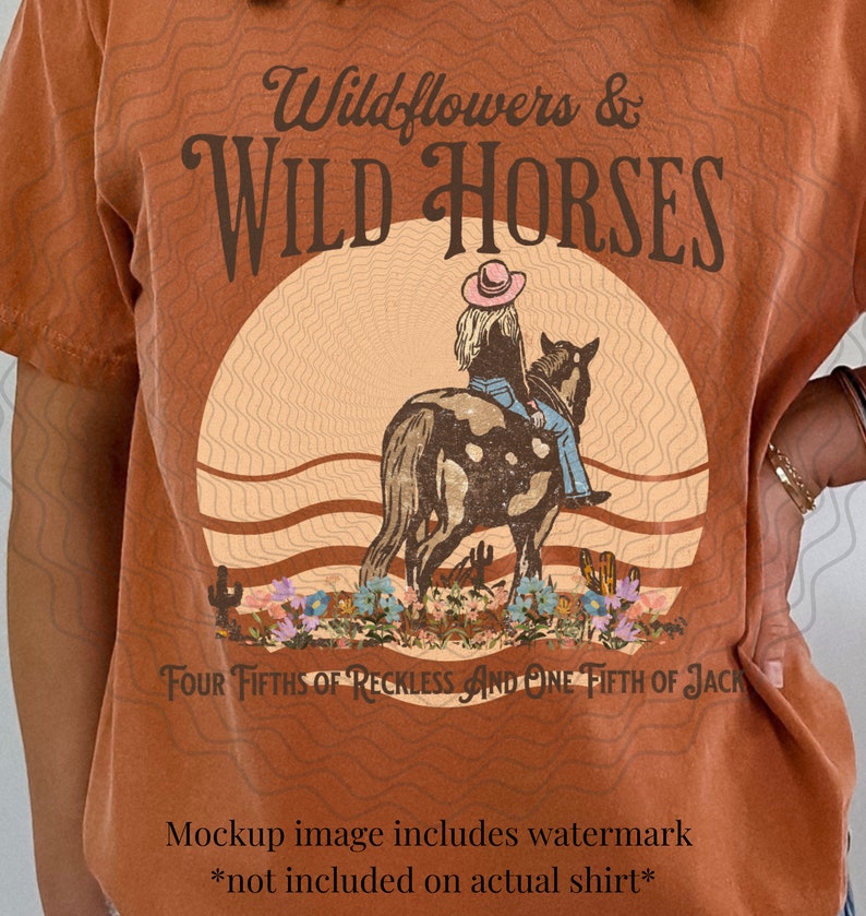 K&ouml;nnte beinhalten: Ein rostfarbenes T-Shirt mit einem Grafikdesign einer Frau, die auf einem Pferd vor einem Sonnenuntergang reitet. Der Text "Wildflowers & WILD HORSES" befindet sich oben im Design. Der Text "FOUR FIFTHS OF RECKLESS AND ONE FIFTH OF JACK" befindet sich unten im Design.