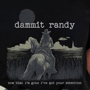 May include: Black t-shirt with a distressed graphic of a person riding a horse, wearing a cowboy hat. The text "dammit randy" is above the image, and the text "now that I'm gone I've got your attention" is below.
