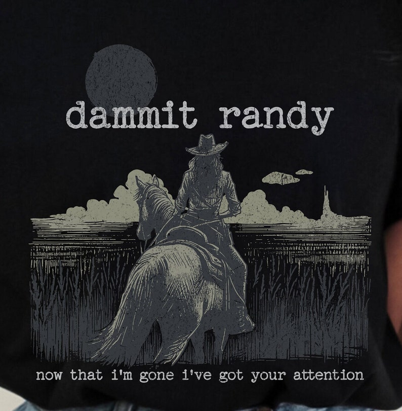 May include: Black t-shirt with a distressed graphic of a person riding a horse, wearing a cowboy hat. The text "dammit randy" is above the image, and the text "now that I'm gone I've got your attention" is below.