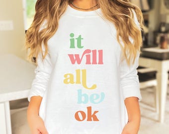 May include: White long-sleeve sweatshirt with the phrase "it will all be ok" in colorful, stacked letters. The text is in green, pink, yellow, light blue, and orange. The sweatshirt is a casual, comfortable style.