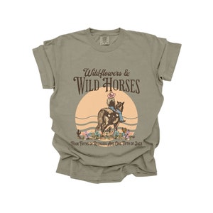 K&ouml;nnte beinhalten: Ein hellgr&uuml;nes T-Shirt mit einem Aufdruck einer Frau, die auf einem Pferd in einer Sonnenuntergangsszene reitet. Der Text "Wildflowers & WILD HORSES" ist in Braun &uuml;ber dem Aufdruck gedruckt. Der Text "Four Fifths of Reckless and One Fifth of Jack" ist in Braun unter dem Aufdruck gedruckt.