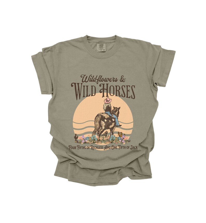 K&ouml;nnte beinhalten: Ein hellgr&uuml;nes T-Shirt mit einem Aufdruck einer Frau, die auf einem Pferd in einer Sonnenuntergangsszene reitet. Der Text "Wildflowers & WILD HORSES" ist in Braun &uuml;ber dem Aufdruck gedruckt. Der Text "Four Fifths of Reckless and One Fifth of Jack" ist in Braun unter dem Aufdruck gedruckt.