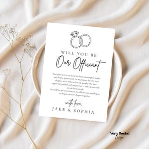 Peut inclure: Une carte blanche avec le texte "WILL YOU BE Our Officiant" et un dessin d'alliance. La carte comprend une demande pour officier un mariage, signée "Jake & Sophia". La carte est sur une assiette blanche avec un fond beige.