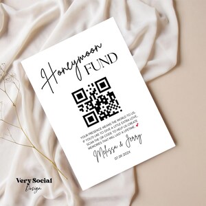 May include: A white card with the words "Honeymoon Fund" in elegant script and a QR code. Below, text reads "Your presence means the world to us..." followed by names and a date. The card is on a cream-colored fabric.
