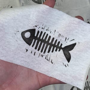 May include: A white fabric square with a black fish skeleton design. The fish skeleton is a detailed illustration with a round eye and a tail fin. The fabric is held in a hand, with other similar prints visible in the background.