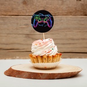 May include: A white cupcake with pink frosting and colorful sprinkles topped with a round black and white cake topper featuring a neon green, blue, and purple video game controller.