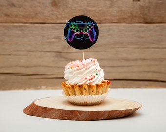 May include: A white cupcake with pink frosting and colorful sprinkles topped with a round black and white cake topper featuring a neon green, blue, and purple video game controller.