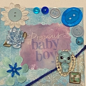 May include: A handmade baby-themed canvas art piece. The canvas is decorated with blue and white embellishments, including buttons, flowers, and a small bear figurine. The text "Precious baby boy" is visible.