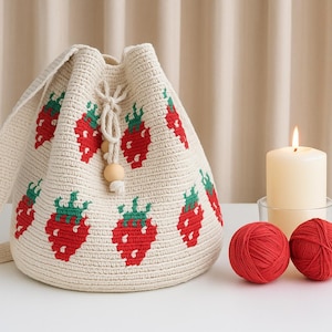 May include: Cream crochet bucket bag featuring a red strawberry pattern with green leaves. The bag has a drawstring closure with wooden beads and a shoulder strap. Two red yarn balls and a lit candle are also in the image.