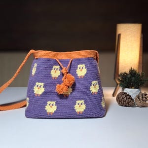 May include: A handmade purple crochet bag with a repeating pattern of yellow chicks. The bag has an orange strap and trim, with a matching tassel. The bag is sitting on a white surface.