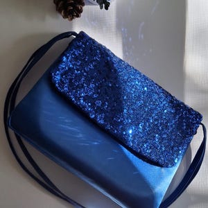May include: A blue sequined clutch bag with a long strap. The bag has a flap closure and is made of a shiny fabric.