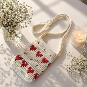 May include: Cream-colored crochet bag with a red heart pattern. The bag has a long strap and is sitting on a white surface. A lit candle and a vase of flowers are also in the image.