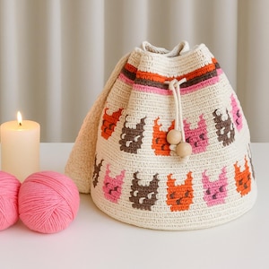 May include: A cream crochet bucket bag with a drawstring closure, patterned with brown, orange, and pink shapes. Two pink yarn balls and a lit candle are in the foreground. The bag is likely handmade.
