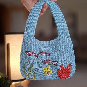 Könnte beinhalten: Eine hellblaue Handtasche, die mit kleinen Perlen besetzt ist und eine Unterwasserszene mit zwei kleinen Fischen, grünem Seegras, einem gelben Seestern und einer roten Korallenformation zeigt. Die Tasche hat einen integrierten Griff.