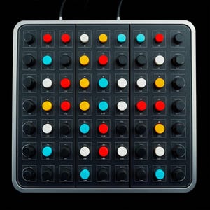 以下が含まれることがあります： ボタンとノブのグリッドを備えた黒い電子音楽制作デバイス。ボタンの色は赤、黄、白、青など様々です。デバイスはシルバーフレームと黒いケーブルを備えています。