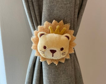 May include: A plush lion curtain tieback in shades of tan, beige, and brown. The lion has a golden crown and is embroidered with facial features. The tieback is holding a gray curtain.