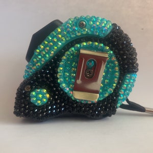 May include: A black and turquoise measuring tape with rhinestones. The tape has a metal clip and a black strap.