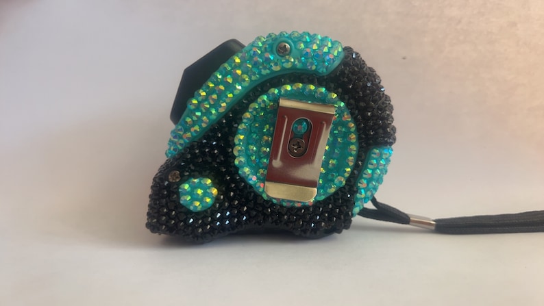 May include: A black and turquoise measuring tape with rhinestones. The tape has a metal clip and a black strap.