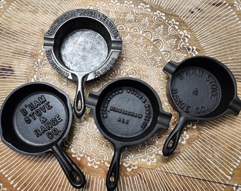 May include: Four cast iron ashtrays with handles. Three are black and one is silver. The ashtrays are all round and have the text "Birmingham Stove & Range Co." and "Alabama" on them.