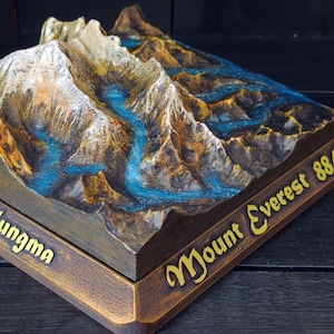 Puede incluir: Un modelo de madera del Monte Everest con una representación detallada del terreno de la montaña y un río azul que fluye por el lado. La base del modelo está inscrita con "Mount Everest 8848m" en letras doradas.