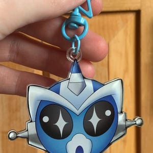 May include: A blue and white cartoon character keyring with a star-shaped clip. The character has large black eyes with star-shaped highlights, a surprised expression, and a robotic design. The keyring is held by a hand.