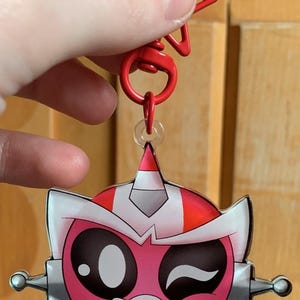 May include: A pink and red cartoon character keychain with a winking expression. The keychain has a red star-shaped clasp and a silver-colored metal accent. The character has large black eyes and a white and silver helmet.