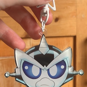 May include: A cartoon-style keyring featuring a robot-like character with a white and silver design and large blue eyes. The keyring has a white star-shaped top and a metal clasp.