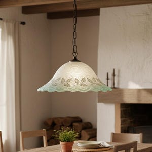 May include: A pendant light fixture with a white, scalloped shade featuring a leaf design in a light green hue. The light hangs from a dark chain and is suspended above a wooden dining table. The room has a fireplace and a small potted plant.