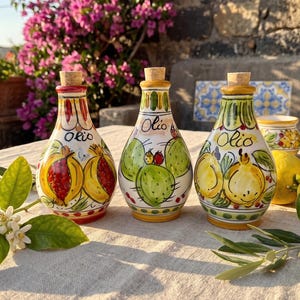 Puede incluir: Tres aceiteras de cerámica con tapones de corcho, cada una con diseños pintados a mano. La de la izquierda muestra granadas, la central higos chumbos y la de la derecha limones. Cada botella está etiquetada con la palabra Olio en cursiva.