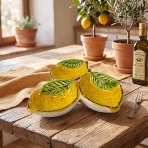 May include: A three-section ceramic serving dish shaped like three lemons joined together, featuring bright yellow textured interiors, green leaves on the rims, and white exteriors, accompanied by a small silver metal fork.