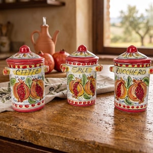 May include: Three ceramic canisters with lids, each featuring hand-painted pomegranate illustrations and red trim. The canisters are labeled CAFFE, SALE, and ZUCCHERO in black script. Each container has two small side handles and a pointed lid top.
