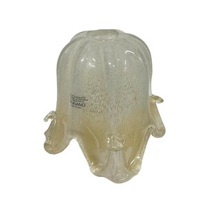 Op de afbeelding: Een helder, klokvormig glazen object met een gespikkelde, gebroken witte en licht amberkleurige kleur. Het glas heeft een getextureerd uiterlijk en een uitlopende, geschulpte rand. De basis heeft kleine, afgeronde uitsteeksels. De tekst "MURANO" is zichtbaar.