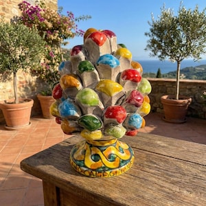 Pode incluir: Uma escultura de cerâmica colorida que se assemelha a uma pinha, com uma base com padrões amarelos e azuis. A escultura tem uma infinidade de formas arredondadas de cores vivas em vermelho, amarelo, verde e azul. Está sobre uma mesa de madeira.
