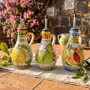 Pode incluir: Três galheteiras de cerâmica com bico de metal, cada uma com desenhos pintados à mão de um limão amarelo, um cacto verde e uma romã vermelha. Cada galheteira apresenta a palavra Olio escrita em letra cursiva na parte da frente.