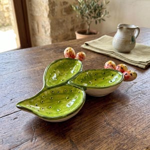 May include: A ceramic dish shaped like a prickly pear cactus pad with three concave sections. The interior is bright green with small dark dots, while the exterior is off-white. Attached to the edges are small, rounded, orange-red fruits.
