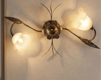 Könnte beinhalten: Wandleuchte mit floralem Design. Die Leuchte hat eine bronzefarbene Oberfläche und verfügt über zwei tulpenförmige Glasschirme. Das zentrale Element ist eine Blume mit zarten Blütenblättern, und die Leuchte enthält Blätter.