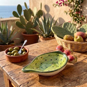Puede incluir: Un plato de cerámica con forma de pala de chumbera, con esmalte verde claro y puntos verde oscuro. A su lado hay un cuenco de terracota con aceitunas verdes y un tenedor de plata, además de una cesta de mimbre llena de higos chumbos.