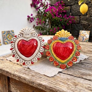 Può includere: Due ornamenti decorativi da parete a forma di cuore con un cuore centrale rosso lucido circondato da cornici ornate e scolpite. Una cornice è color crema con accenti rossi, mentre l'altra è verde e gialla con dettagli floreali rossi.