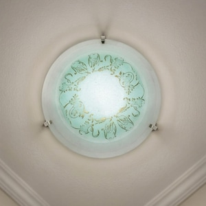 May include: A circular, frosted glass ceiling light with a teal and gold floral pattern. The light is fixed with silver metal brackets. The fixture is centred on a white ceiling.