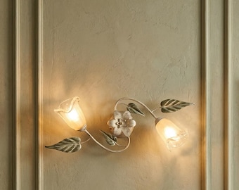 Könnte beinhalten: Dekorative Wandleuchte mit floralem Design. Sie verfügt über zwei tulpenförmige Lampen mit einer zentralen Blume und Blattdetails. Der Metallrahmen hat eine cremefarbene, antike Oberfläche, und die Lampen sind beleuchtet.