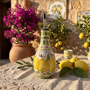 May include: A ceramic bottle with a tapered neck and a metal pour spout, decorated with hand-painted yellow lemons, green leaves, and the word Limoncello in cursive script. The bottle features green and red accents on a cream-coloured background.