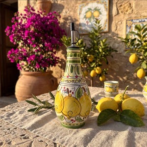 Puede incluir: Una botella de cerámica con cuello cónico y vertedor de metal, decorada con limones amarillos pintados a mano, hojas verdes y la palabra Limoncello en letra cursiva. La botella presenta detalles en verde y rojo sobre un fondo color crema.