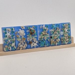 Könnte beinhalten: Sechs blaue Dominosteine aus Kunstharz, jeweils mit Muscheln, Seesternen und kleinen grünen Perlen. Die Dominosteine sind auf einer hellen Holzbasis angeordnet und erzeugen ein maritimes Thema. Ein einzigartiger Dekorationsartikel.