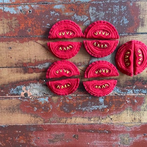 May include: Six felt tomato slices, each with a red felt exterior and yellow felt stitching to represent seeds. The slices are arranged in two rows of three on a wooden surface.