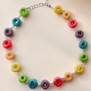 May include: A colorful necklace featuring a series of rainbow-colored, circular beads strung on a silver chain. The beads are in shades of red, orange, yellow, green, blue, and purple. The necklace is displayed on a light beige background.