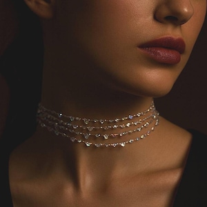May include: A woman wearing a multi-strand necklace with sparkling diamond-like accents. The necklace is composed of several delicate strands, each adorned with small, glittering stones. The woman is wearing a black top, and her skin has a warm, golden tone.