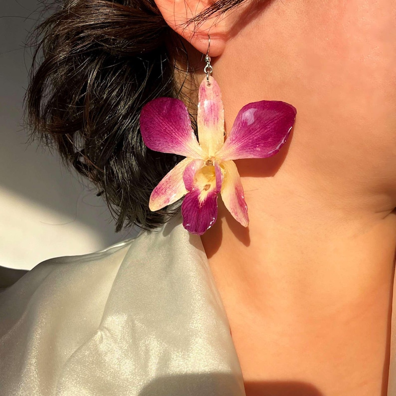May include: A pair of earrings featuring a purple and yellow orchid flower. The earrings are made of resin and have a silver hook.