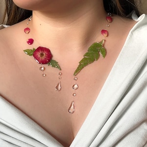 May include: A handmade necklace featuring a red flower and green leaves, with clear teardrop-shaped pendants. The necklace also includes small, clear beads and pink flower petals. The design is set against a light background.
